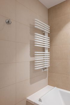 Corner of bathroom in the center with a white heating radiator on the wall for drying towels. The walls of the room are lined with beige tiles of different colors. Pure white acrylic bathtub