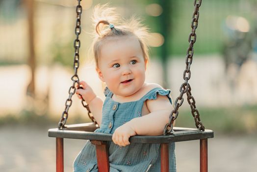 Toddler baby girl on a swing on the warm summer evening. Mother is swinging her young daughter on a sunny playground.