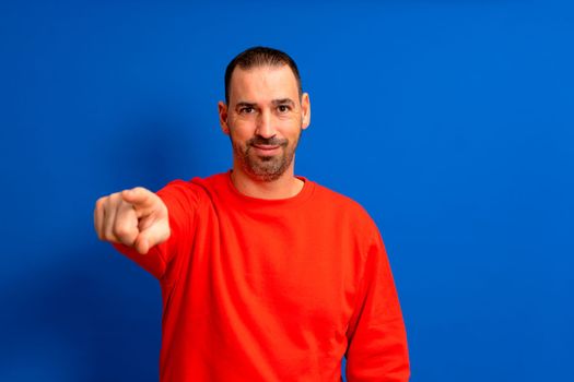 Portrait of a hispanic man with a beard wearing a red pullover pointing forward, looking at the camera. Half-length portrait on blue background.