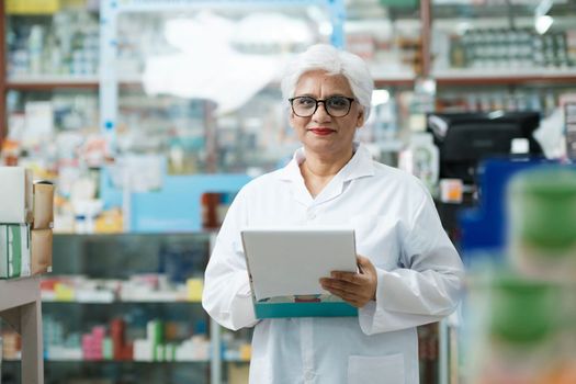 Female smart senior pharmacist in professional white gown looking at camera and checking stock inventory in modern pharmacy, drugstore indoor holding cli. Pharmacy, medicine, and healthcare concept.