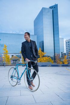 Young smiling stylish African American businessman in suit going to the office by bicycle in front of financial district. Business and transport concept. High quality vertical photo
