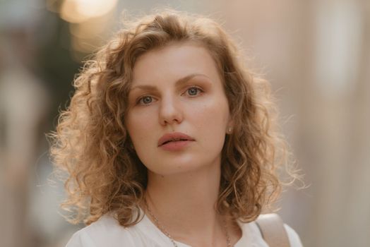 A headshot photo of a woman with curly hair in an old European town. A lady in a blurry background in the evening.