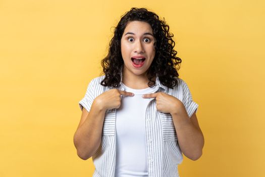 Portrait of woman with dark wavy hair pointing herself, looking with surprised expression, expressing shock, unbelievable success. Indoor studio shot isolated on yellow background.