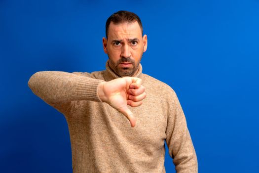 Bearded latino man wearing brown turtleneck looking unhappy and angry showing rejection and negative thumbs down gesture. Bad expression.