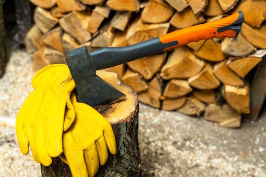 an iron axe stuck in a wooden deck and protective yellow leather gloves against the background of split firewood. Firewood harvesting, preparation for the heating season is a professional tool. An ax is stuck in a wooden deck for chopping firewood