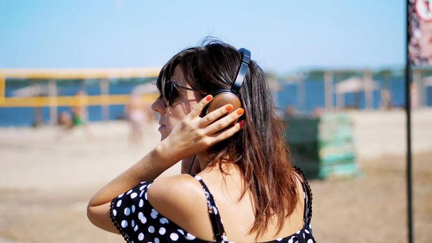 portrait, beautiful girl in sun glasses wearing big black headphones, listening to music from smartphone, on beach, on hot summer day, against background of beach volleyball. High quality photo