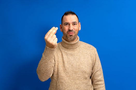 Handsome hispanic man with beard wearing brown turtleneck sweater over blue isolated background doing italian gesture with hand and fingers confident expression