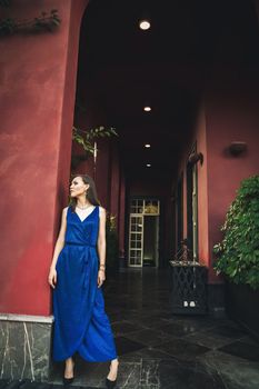 Fashion luxury vertical portrait of young thin woman in blue dress next to rich red big house, copy space.