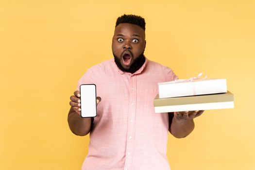 Portrait of shocked astonished man in pink shirt holding present box and smatrphone with blank screen for promotion, looking at camera with big eyes. Indoor studio shot isolated on yellow background.