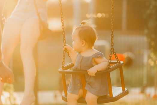 Toddler baby girl on a swing on the warm summer evening. Mother is swinging her young daughter on a sunny playground.