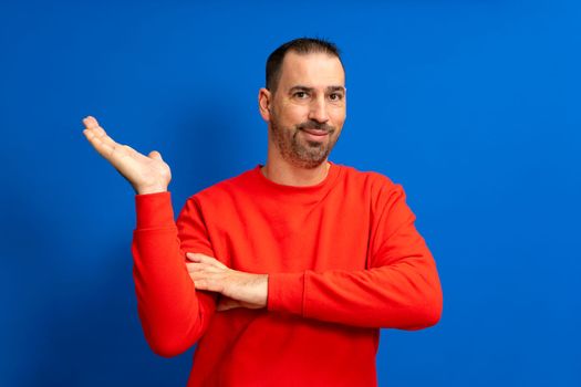 Latino man with beard and red sweater offering something on the palm of his hand, isolated on blue background