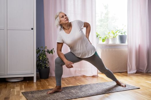 Domestic yoga practice. Positive mature lady standing in yoga pose, keeping balanced during covid lockdown at home. Happy senior lady in sportswear exercising 