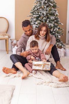 A young stylish multi-racial family with a little boy is sitting on a knitted blanket near the Christmas tree, holding a wooden box with Christmas toys. Vertical