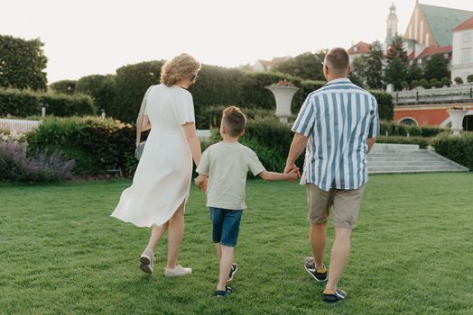 A photo from the back of a happy family is strolling the garden of the palace in an old European town. A smiling father, mother, and son are holding hands and having fun at sunset