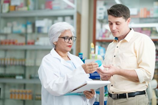 Female senior pharmacist at the drugstore wearing white gown talking, giving advice, explaining, suggesting, and recommending to client or patient about the prescription and medications. Medicine and healthcare concept.