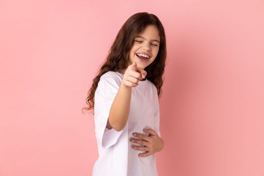 Portrait of positive optimistic joyful little girl wearing white T-shirt laughing out loud holding belly and pointing finger on you, mockery. Indoor studio shot isolated on pink background.