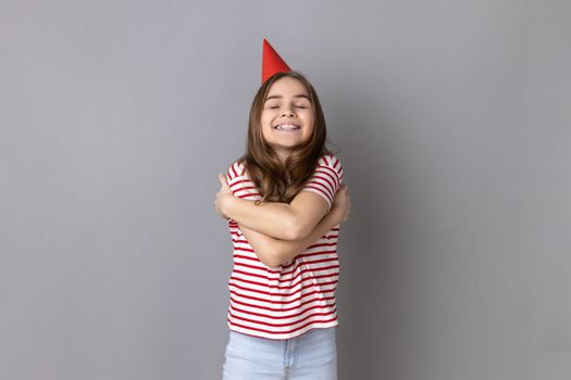 Little girl wearing striped T-shirt and party cone hugging oneself with expression of pleasure and love, celebrating birthday, congratulating herself. Indoor studio shot isolated on gray background.