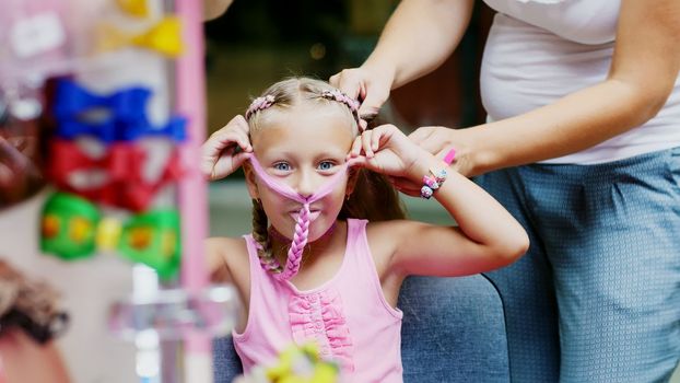 Beautiful blond girl, of seven years old, braided two pigtails, do a hairstyle with pink locks of hair in a beauty salon, a hairdresser's salon, in front of a large mirror. a little princess. High quality photo