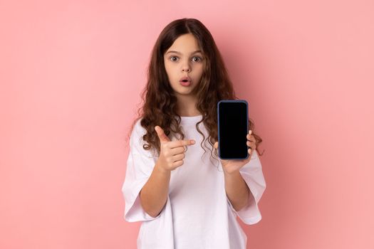 Portrait of shocked surprised little girl wearing white T-shirt showing smart phone with black blank screen, pointing finger at display. Indoor studio shot isolated on pink background.