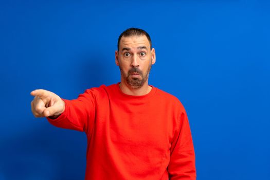 Bearded latino man in a red pullover pointing finger at the camera isolated over blue background