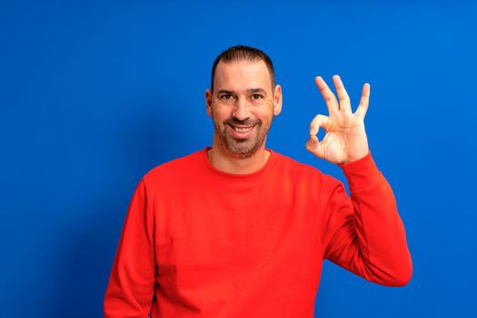 Handsome smiling happy bearded latino man in his 40s wearing red sweatshirt making ok gesture isolated over blue studio background. People lifestyle concept