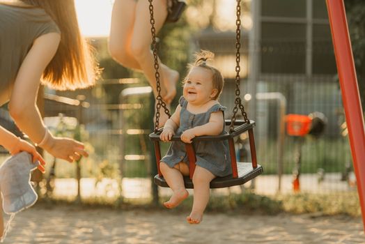 Toddler baby girl on a swing on the warm summer evening. Mother is swinging her young daughter on a sunny playground.