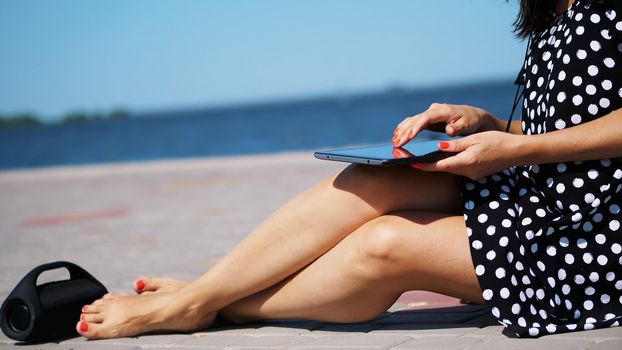 close-up, girl works on a tablet, female hands, legs with bright red manicure, nails, on the beach, on a hot summer day, listening to music with bluetooth portable speaker. High quality photo