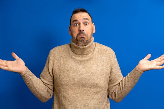 Hesitating bewildered bearded fashionanble man full of doubts, must decide or choose between two proposals, pursed lips, has indecisive expression, shows uncertainty, gestures against blue background