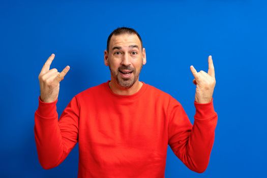 Portrait of attractive hispanic man wearing red jumper over isolated blue background screaming with crazy expression doing rock symbol with hands up. Music star,heavy concept.