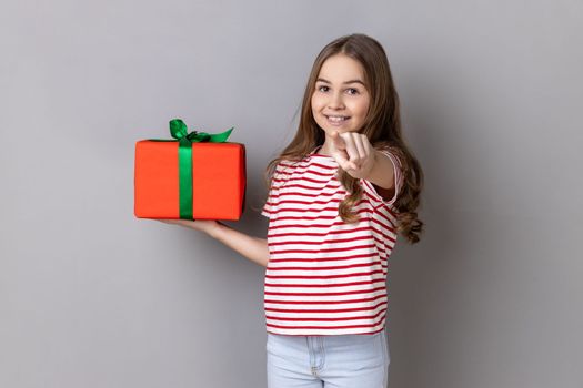Hey, you are winner. Portrait of delighted little girl wearing striped T-shirt holding present box and pointing finger to camera with excited expression. Indoor studio shot isolated on gray background