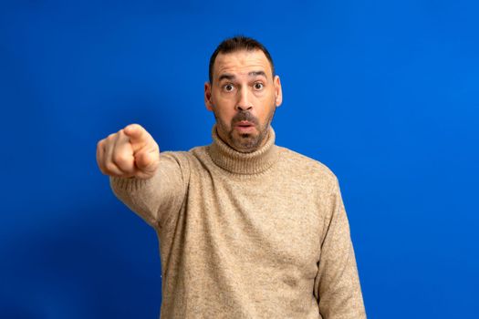 Portrait of a bearded hispanic man in brown turtleneck pointing finger at camera isolated over blue background.
