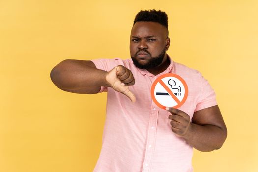 Portrait of serious young adult bearded man wearing pink shirt holding no smoking sign, showing thumb down gesture, looking at camera. Indoor studio shot isolated on yellow background.