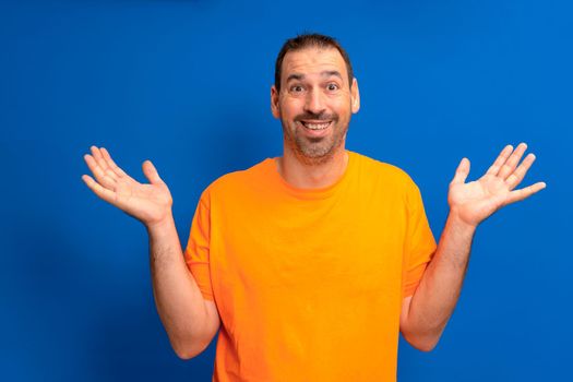 Portrait of satisfied carefree impressed guy wearing orange t-shirt raises palms up and shouts yes isolated over blue tinted background