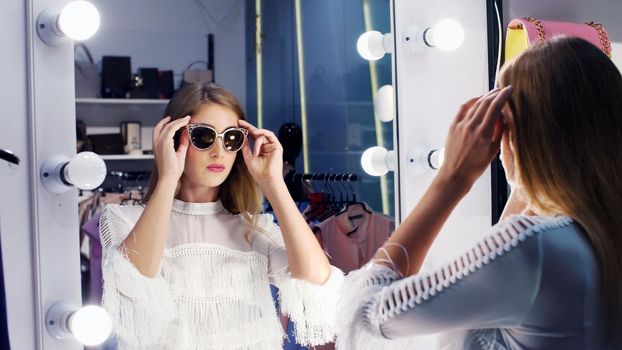Sexy girl, tall, beautiful blond woman trying on sunglasses in a stylish store, boutique and admiring herself in front of a mirror, smiling. slow motion. High quality photo