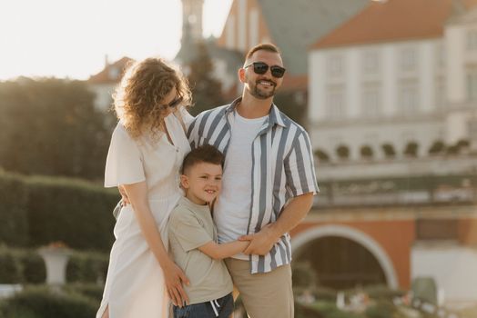 Father, mother and son are hugging in an old European town. Happy family in the evening. Dad is smiling at his family in the background of the palace at sunset.