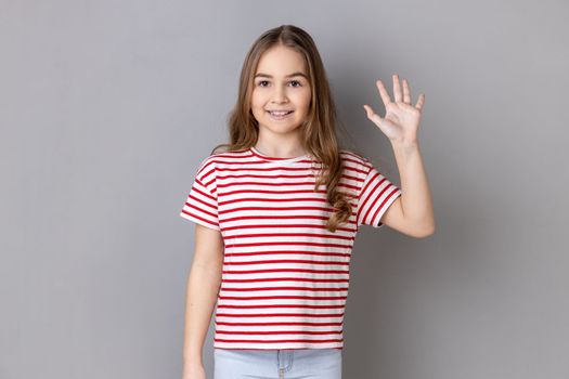Portrait of adorable cheerful little girl wearing striped T-shirt standing waving hand, looking at camera with engaging toothy smile. Indoor studio shot isolated on gray background.