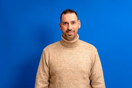Isolated photo of handsome man with beard, mustache and trendy hairstyle, wears brown turtleneck sweater, has joyful expression while listening to his interlocutor, poses in studio blue background