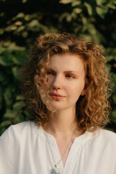 A woman with curly hair is posing in the park at the sunset. A lady with green leaves in the background.