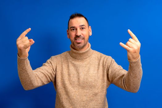 Bearded latino man wearing brown turtleneck over isolated blue background screaming with crazy expression doing rock symbol with hands up. Music star. Heavy concept.