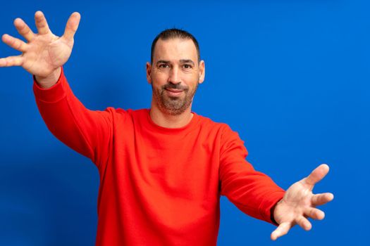 Portrait of a bearded latino man in a red jumper with arms outstretched in welcome isolated over blue studio background