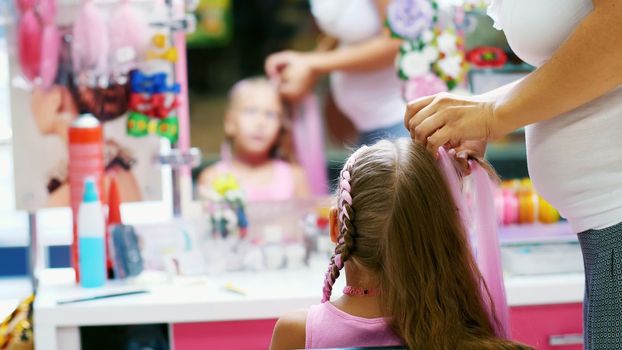 Beautiful blond girl, of seven years old, braided two pigtails, do a hairstyle with pink locks of hair in a beauty salon, a hairdresser's salon, in front of a large mirror. a little princess. High quality photo