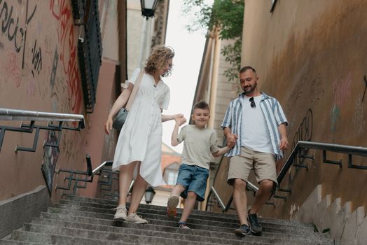 A full-length photo of a family which is going down the stairs in an old European town. A happy father, mother, and son are holding hands and having fun in the evening.