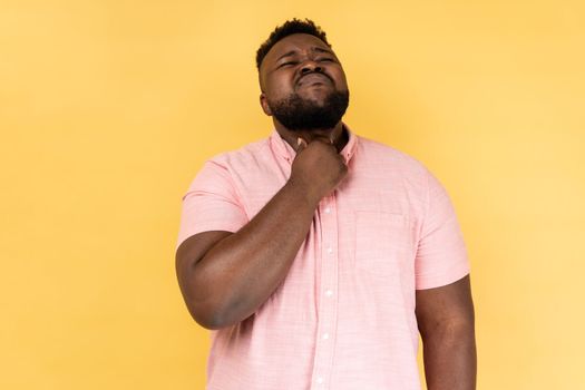 Throat pain. Portrait of sick man wearing pink shirt standing and touching his painful throat, frowning his face, feels terrible pain. Indoor studio shot isolated on yellow background.