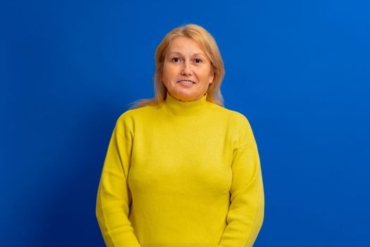 Beautiful blonde woman in yellow sweater looking to side to side with smile on face isolated over blue background, natural expression. Laughing confident.