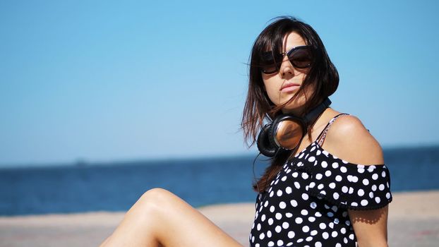 portrait, beautiful brunette, woman in sun glasses, and headphones, sitting on the beach, against the sea. wind waves her hair. High quality photo