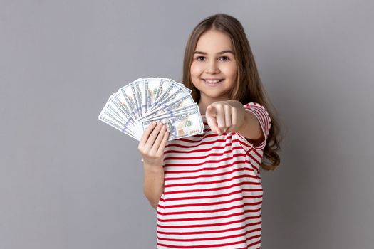 Portrait of delighted little girl wearing striped showing at fan of dollar banknotes, showing big money and pointing at camera. T-shirt Indoor studio shot isolated on gray background.