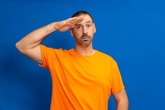 A man in greeting pose and serious face. A hispanic man with a beard wearing orange t-shirt isolated on blue background.