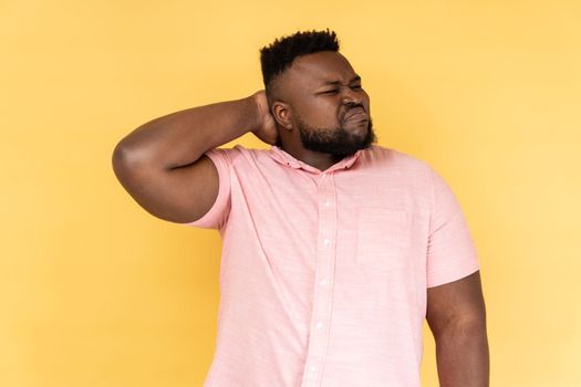 Portrait of worried anxious bearded man wearing pink shirt touching neck, feeling acute pain moving and turning head, suffering spine problems. Indoor studio shot isolated on yellow background.