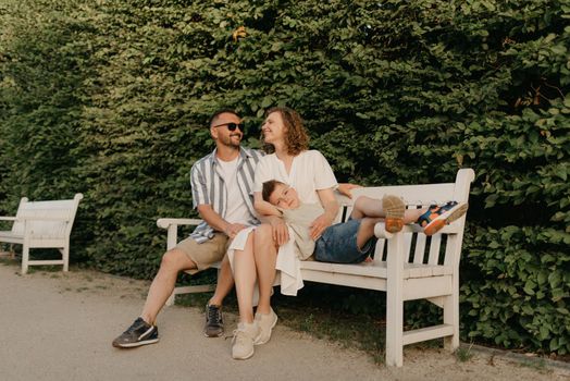 Father, mother, and son are sitting on the bench in the garden of an old European town. Laughing family in the park in summer at sunset.