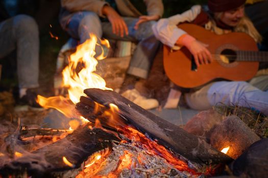 Unrecognizable friends having fun playing music and enjoying bonfire camping in nature at night. Group of people chilling at fire in the evening, camping in the forest near lake. High quality photo